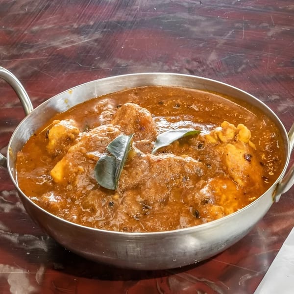 A metal karahi bowl filled with rich, orange-brown Indian curry, garnished with green leaves.