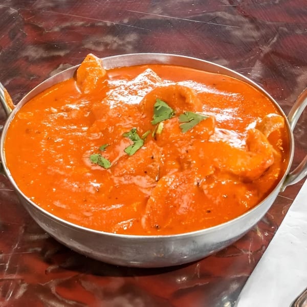 A metal bowl of vibrant orange Indian butter chicken, garnished with fresh cilantro, on a dark table.